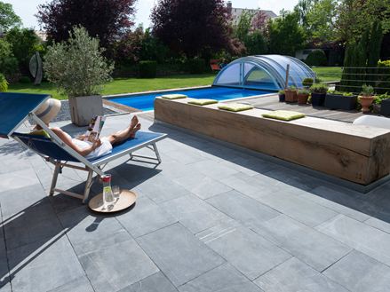 Vrouw ontspant op een ligstoel op een terras met kalkstenen platen, op de achtergrond een tuin met zwembad
