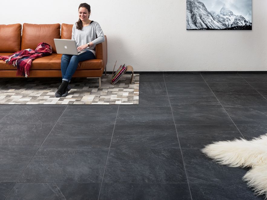 Woonkamer met tegels in leisteenlook, een vrouw zit op een bruine bank met een laptop