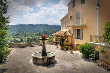 Een fontein op een plein naast een landhuis, bergen op de achtergrond