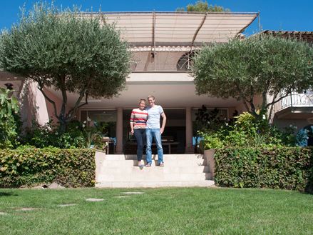 Staand koppel voor een hotel met travertin vloer, een gazon op de voorgrond en bomen links en rechts