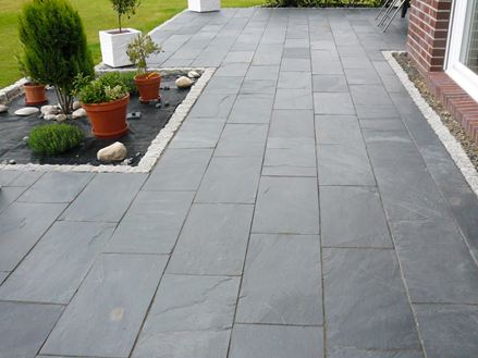 Terras met potplanten en zwarte rustieke leisteen als vloerbedekking, rechts zie je een bakstenen huismuur