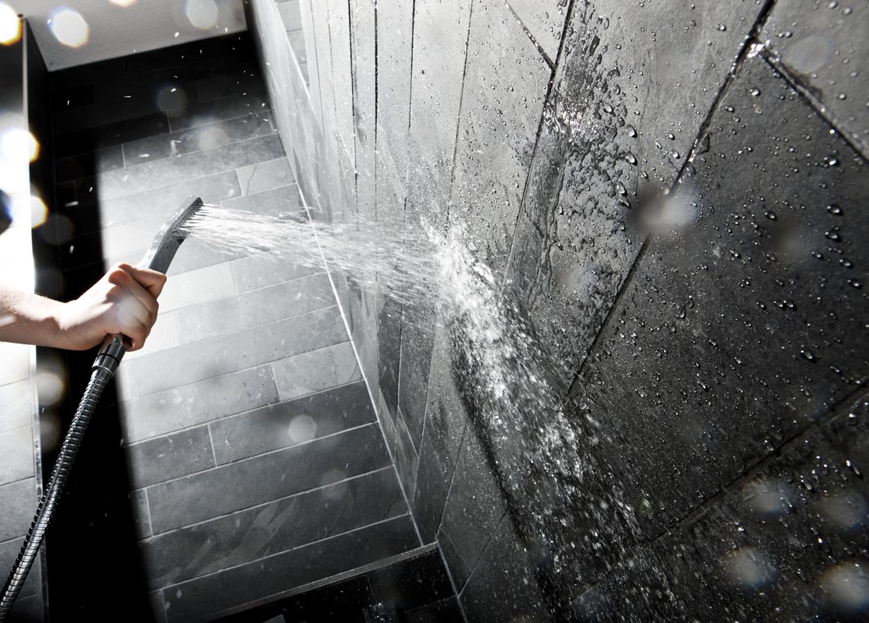 Hand houdt een douchekop tegen een muur met donkere stenen tegels